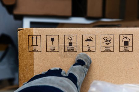 Photo for In a warehouse, a male worker wearing gloves points to cardboard boxes adorned with handling instructions in both English and Chinese, including This Side Up, Fragile, Stacking Limit, Keep Dry, Do Not - Royalty Free Image