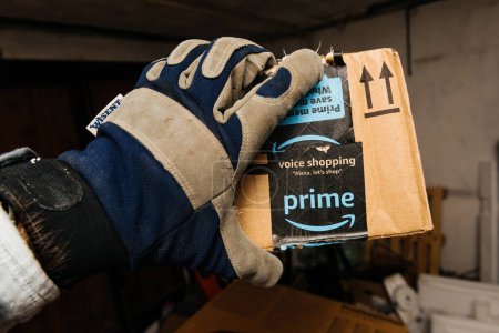 Foto de Bremen, Alemania - 10 de diciembre de 2023: En un almacén grande y débilmente iluminado, una mano masculina que lleva un guante sostiene un paquete de cartón Amazon Prime, con entrega rápida y gratuita tan rápido como hoy, con el Todo - Imagen libre de derechos