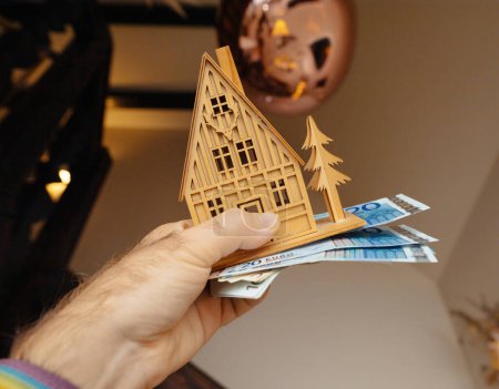 Téléchargez les photos : Une main de propriétaire tient délicatement une petite maison en bois jouet aux côtés d'une pile d'argent, symbolisant les revenus d'investissement immobilier et de location, situé dans un intérieur de luxe moderne - en image libre de droit