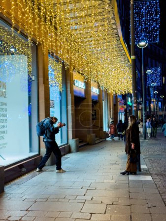 Foto de Estrasburgo, Francia - 25 de diciembre de 2023: Un fotógrafo masculino captura a una mujer elegante en medio de las encantadoras decoraciones navideñas cerca de Galerías Lafayette en el centro de Estrasburgo, creando una escena atemporal - Imagen libre de derechos