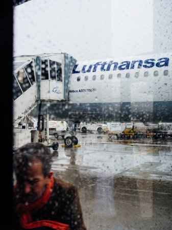 Téléchargez les photos : Francfort, Allemagne - 4 mai 2019 : Une personne portant une écharpe rouge observée à travers une fenêtre mouchetée par la pluie sur fond d'Airbus A321 de Lufthansa. - en image libre de droit