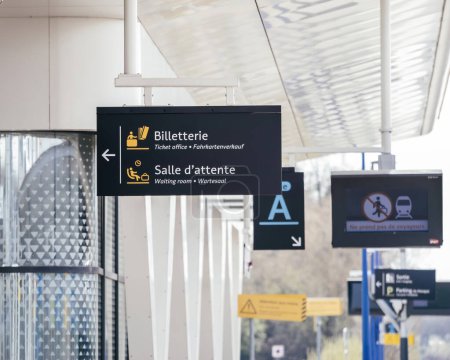 Multilingual sign at a French train station indicating directions to the ticket office and waiting room, with pictograms for clear guidance.