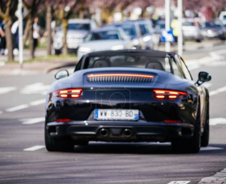 Foto de Haguenau, Francia - 20 de marzo de 2024: Vista borrosa desenfocada de un Porsche 911 Carrera GTS negro en las calles de Haguenau, sus luces traseras únicas brillando mientras navega a través del tráfico de la ciudad - Imagen libre de derechos