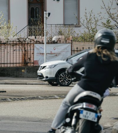Foto de Haguenau, Francia - 20 de marzo de 2024: Una mujer monta con confianza una motocicleta en una bulliciosa calle de la ciudad rodeada de edificios y tráfico. - Imagen libre de derechos