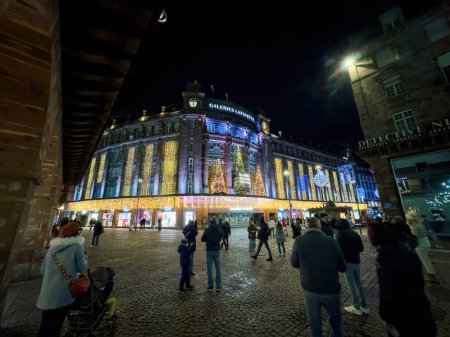 Foto de Estrasburgo, Francia - 31 de diciembre de 2023: En Estrasburgo, las majestuosas Galerías Lafayette deslumbra por la noche en el último día de diciembre. transeúntes, en medio de decoraciones festivas, capturar fotos, añadir a la - Imagen libre de derechos