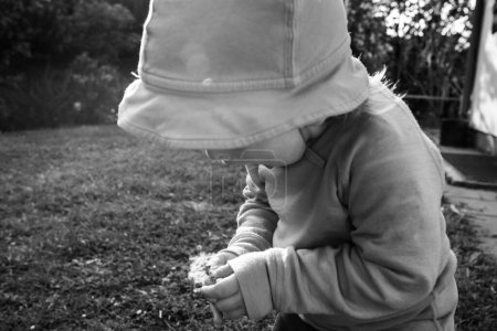 A black and white image captures a boy about to blow seeds from a magnificent dandelion in a garden.