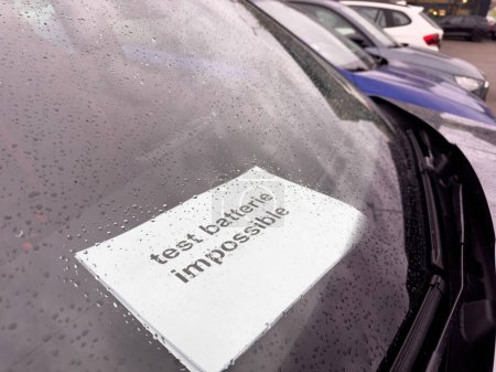A note saying test batterie impossible in french language placed under the windshield wiper of a car in a showroom parking