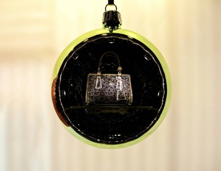 Un sac à main en cuir traditionnel, annoncé à l'intérieur d'un globe de Noël allumé, symbolise un cadeau désiré pour les femmes du monde entier pendant la période des fêtes, alliant élégance et charme festif.