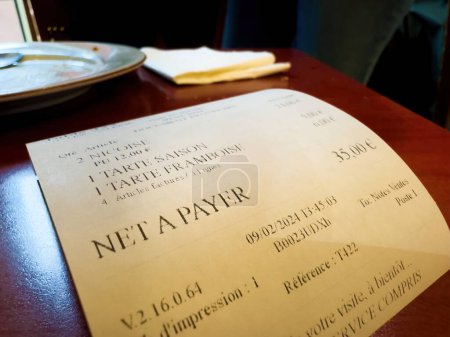 Foto de Imagen de primer plano de un recibo de restaurante colocado sobre una mesa de madera en un café francés. La factura detalla diversos alimentos y la cantidad total adeudada en euros. Una placa terminada se ve en el fondo - Imagen libre de derechos