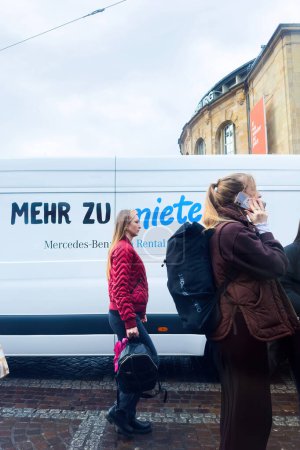 Téléchargez les photos : Fribourg, Allemagne - 5 mars 2024 : Une femme marche près du Théâtre Fribourg bâtiment sur Bertoldstrasse, parler au téléphone, tandis qu'une Mercedes-Benz van de location passe en arrière-plan, se mélangeant avec la - en image libre de droit