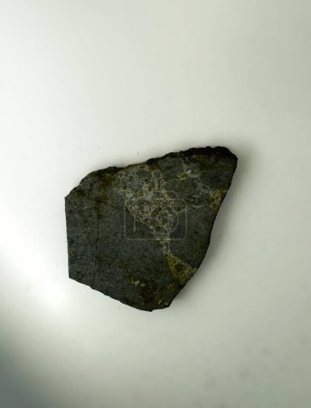 A dark rock fragment with a rough texture lies on a white surface. The rock appears to have natural fractures and variations in color, showcasing its unique geological features
