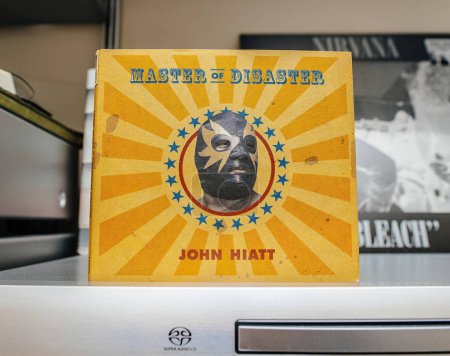 Photo for The SACD album Master of Disaster by John Hiatt is prominently displayed on a shelf. The bright yellow cover with a masked figure stands out against a backdrop of other records. - Royalty Free Image