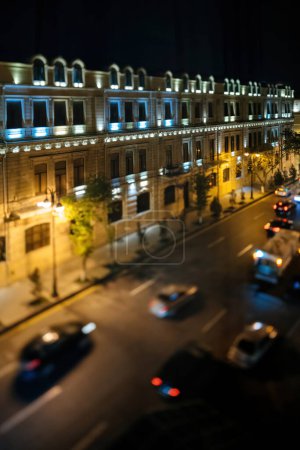 Una vista nocturna de un edificio bellamente iluminado en Bakú, con un efecto de cambio de inclinación que destaca la arquitectura y los coches borrosos en la carretera de abajo, capturando la vibrante vida nocturna de la ciudad