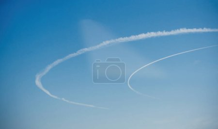 Gebogene Kondensstreifen vor einem klaren blauen Himmel bilden anmutige Bögen, die die Flugbahn eines Flugzeugs in Bewegung hervorheben und eine ruhige und dynamische Luftaufnahme schaffen.