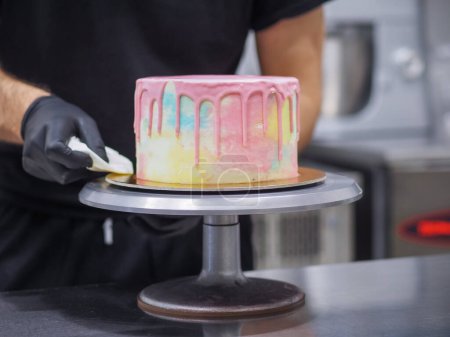 Photo for Pasty chef artisan squeeze piping bag filling mixed with edible pink paint for dripping color grading frosted rainbow pastel cake - Royalty Free Image