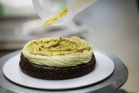 Photo for Pastry chef designer using pistachio cream and sprinkles on layered dark frosted chocolate cake at kitchen lab - Royalty Free Image