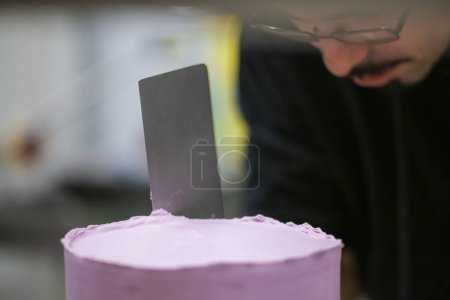 Téléchargez les photos : Chef pâtissier utilisant spatule et grattoir lissage nivellement couches lilas beurre crème gâteau givré - préparation gâteau de mariage - en image libre de droit