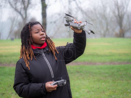 Photo for Cheerful young teenager african american wearing sportswear checking flying a camera drone in a park before use. - Royalty Free Image