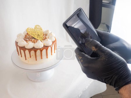 Photo for Chef pastry baker making a reel of a salty caramel frosted dripping cakes with meringues and chocolate heart topper with smarphone photo - Royalty Free Image