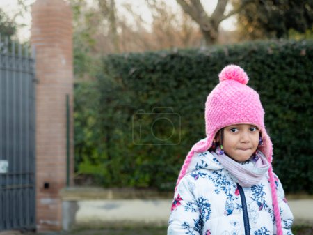 Photo for Cute smiling african american baby girl in white outfit outdoors at sunset wearing a pink beanie knit hat with earflaps - Royalty Free Image