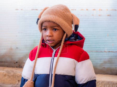 Photo for Portrait of african kid sta ding outdoors in a park a sunny day wearing winter garment and a beanie hat with earflaps - Royalty Free Image