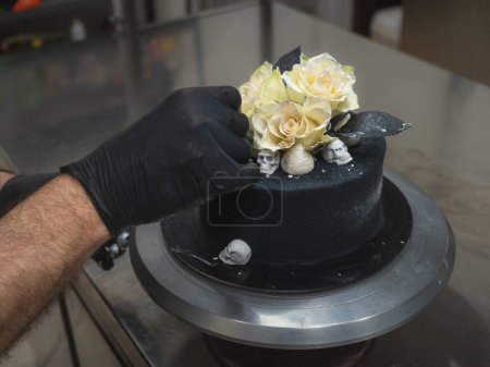 Photo for Pastry chef baker topping birthday black airbrush painted frosted cake, two real roses silver sprayed and edible chocolate skull toppers, silver brush strokes - Royalty Free Image