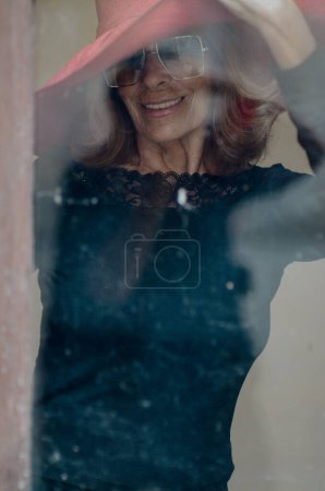 Photo for A woman with a hat on her head stands behind a window indoors while facing the camera, - Royalty Free Image