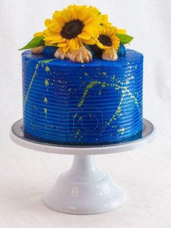 Stunning cake with bright blue icing, adorned with fresh sunflowers and figs, displayed on a white cake stand against a neutral background