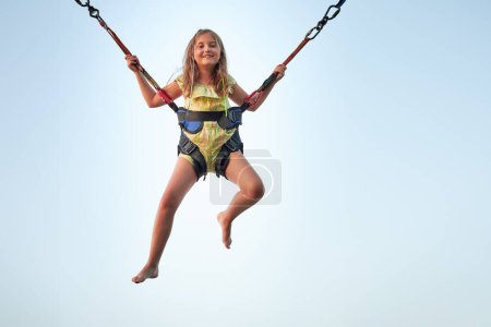 Photo for Bungee jumping at trampoline. Little girl bouncing on bungee jumping in amusement park on summer vacations. Kid flying over blue sky while jumping on trampoline - Royalty Free Image