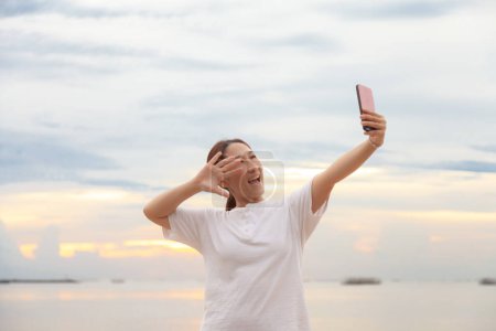 Foto de Las mujeres asiáticas toman fotos y videollamadas en el mar y la playa por la mañana al amanecer con sus teléfonos móviles, teléfonos inteligentes para publicar en Internet en línea mientras viajan de vacaciones.. - Imagen libre de derechos