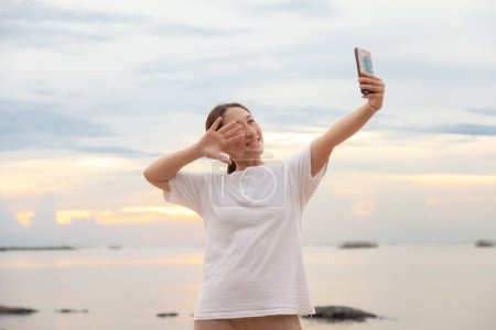 Téléchargez les photos : Les femmes asiatiques prennent des photos et des appels vidéo en mer et à la plage le matin au lever du soleil avec leurs téléphones mobiles, smartphones pour poster sur Internet en ligne pendant leurs vacances. - en image libre de droit