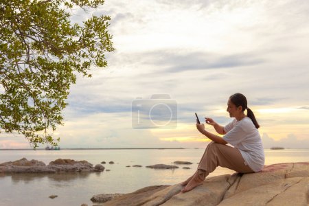 Téléchargez les photos : Les femmes asiatiques prennent des photos et des appels vidéo en mer et à la plage le matin au lever du soleil avec leurs téléphones mobiles, smartphones pour poster sur Internet en ligne pendant leurs vacances. - en image libre de droit