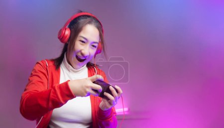 Téléchargez les photos : Heureuse joueuse asiatique avec casque jouant jeu mobile sur le smartphone en ligne. streamers professionnels jeux en vêtements rouges streaming sur le jeu de jeu social avoir du plaisir. Style de lumières Cyber Neon - en image libre de droit