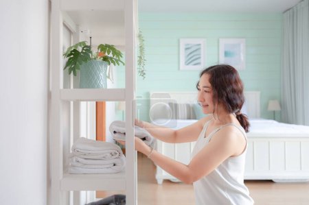 Photo for Positive Asian woman housewife tidying modern home. Happy woman doing domestic chores. Asian housewife folding clothes in the closet. - Royalty Free Image