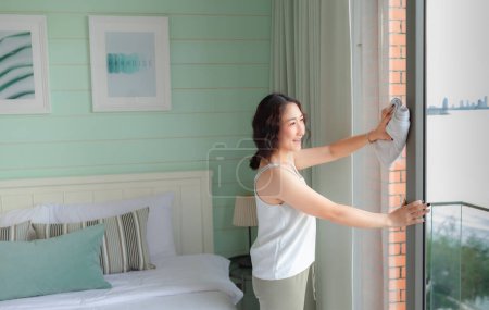 Téléchargez les photos : Femme asiatique positive femme au foyer rangeant maison moderne. Une femme heureuse qui fait des tâches domestiques. Asiatique femme au foyer Essuyage étagère, meubles, lit, miroir, canapé et appareils ménagers. - en image libre de droit