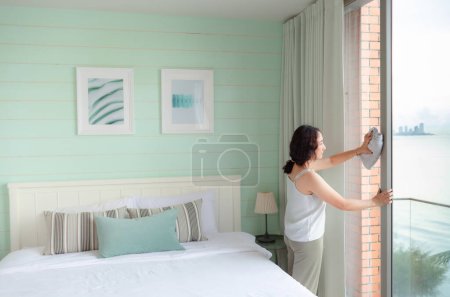 Photo for Positive Asian woman housewife tidying modern home. Happy woman doing domestic chores. Asian housewife Wiping shelf, furniture, bed, mirror, sofa, and home appliances. - Royalty Free Image