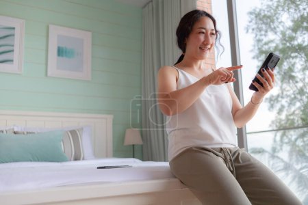 Téléchargez les photos : Heureuse femme asiatique utilisant un téléphone mobile à la maison. Asiatique ménagère vérifier la tenue des médias sociaux avec smartphone. relax femme en utilisant l'application de téléphone mobile jouer la livraison de commande, achats en ligne, appel vidéo - en image libre de droit