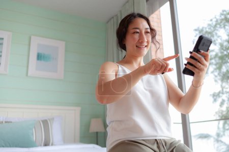Foto de Mujer asiática feliz usando el teléfono móvil en casa. Asiática ama de casa comprobar social media holding con smartphone. relajar a la mujer usando la aplicación de teléfono móvil jugando la entrega de pedidos, compras en línea, videollamada - Imagen libre de derechos