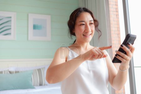 Téléchargez les photos : Heureuse femme asiatique utilisant un téléphone mobile à la maison. Asiatique ménagère vérifier la tenue des médias sociaux avec smartphone. relax femme en utilisant l'application de téléphone mobile jouer la livraison de commande, achats en ligne, appel vidéo - en image libre de droit