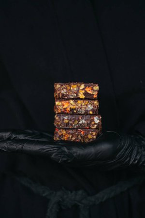 Las manos en guantes negros sostienen barras de chocolate mezcladas con nueces y frutas en el fondo negro. Foto de alta calidad