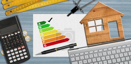 Photo for House energy efficiency rating. Small wooden model house on desk with energy efficiency graph, calculator, folding ruler, drawing compass, pencil and a computer keyboard and copy space. - Royalty Free Image