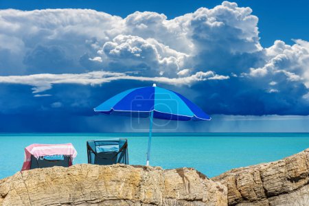Téléchargez les photos : Parapluie de plage bleu et deux chaises longues sur la côte rocheuse avec beau paysage marin avec nuages orageux (cumulonimbus) et pluie torrentielle en arrière-plan. Golfe de La Spezia, Lerici, Italie, Europe. - en image libre de droit