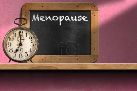Closeup of an old alarm clock above a wooden shelf with a blackboard with text Menopause and copy space. On a Pink wall at seven o'clock.  