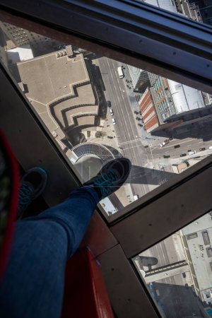 Téléchargez les photos : Debout au-dessus de l'abîme une personne en jeans et baskets se tient au-dessus d'une fenêtre dans le sol à partir de laquelle un panorama de la ville est visible - en image libre de droit