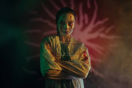 Photo for Young woman portrays bloodthirsty zombie with horror wounds on her face and bloody clothes against dark background with lit. Portrait. Scary image for Halloween. - Royalty Free Image