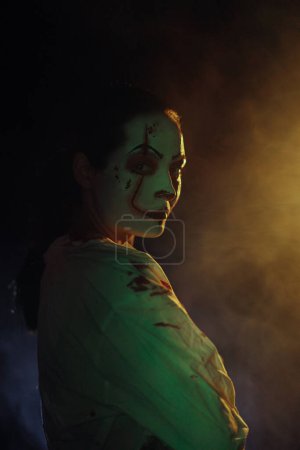 Photo for Young woman portrays bloodthirsty zombie with horror wounds on her face and bloody clothes against dark background with smoke. Scary image for Halloween. Portrait. - Royalty Free Image