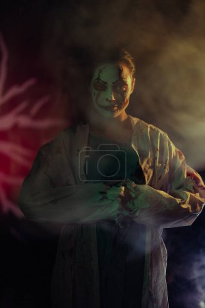 Téléchargez les photos : Jeune femme dépeint zombie sanguinaire avec des blessures horribles sur le visage et des vêtements sanglants sur fond sombre avec de la fumée. Image effrayante pour Halloween. - en image libre de droit