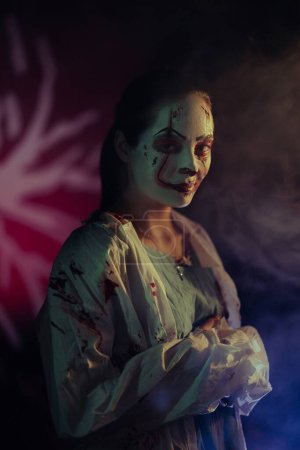 Foto de Mujer joven retrata zombi sediento de sangre con heridas de horror en su cara y ropa sangrienta sobre fondo oscuro con luz. Imagen aterradora para Halloween. - Imagen libre de derechos