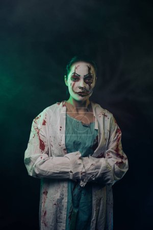 Téléchargez les photos : Jeune femme dépeint zombie sanguinaire avec des blessures horribles sur le visage et des vêtements sanglants sur fond sombre avec éclairé. Image effrayante pour Halloween. - en image libre de droit