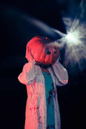 Photo for Young woman holds pumpkin on her head and portrays bloodthirsty zombie with bloody clothes against dark background with backlight. Scary image for Halloween. - Royalty Free Image